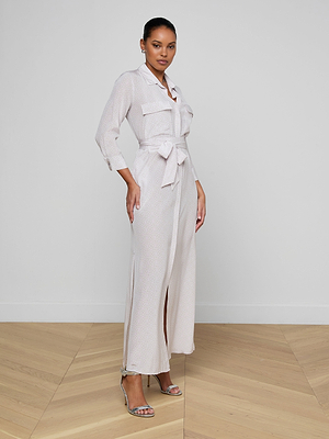 A woman stands indoors wearing the Cameron Silk Shirt Dress, a long, light-colored silk dress with chest pockets and long sleeves. She styles it with high-heeled sandals and a pulled-back hairstyle against a minimal plain background.