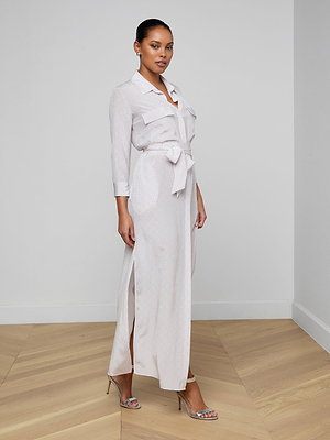 A woman stands in a minimal room wearing the Cameron Silk Shirt Dress—long, light-colored, with long sleeves, front pockets, and side slits—paired with strappy heeled sandals and elegant stud earrings.