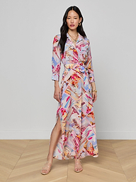 A woman stands wearing the Cameron Silk Shirt Dress, a colorful crepe de chine maxi with long sleeves, a collar, and a side slit. The dress showcases a vibrant abstract pattern in red, pink, blue, and beige tones. She pairs it with sandals, has long black hair, and maintains a neutral expression.
