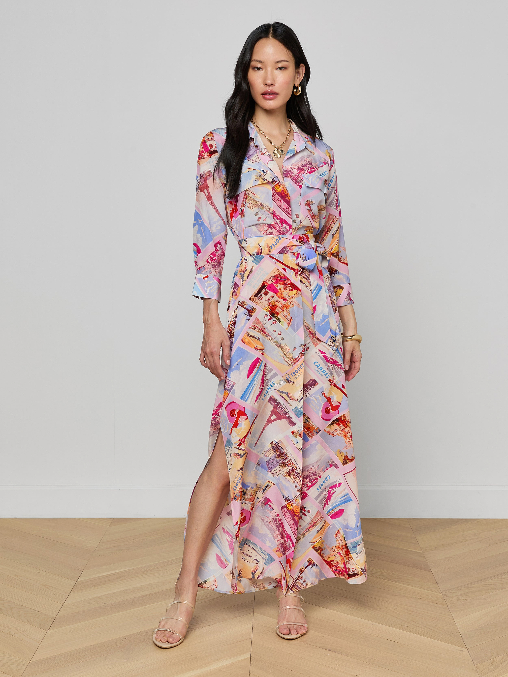 A woman stands wearing the Cameron Silk Shirt Dress, a colorful crepe de chine maxi with long sleeves, a collar, and a side slit. The dress showcases a vibrant abstract pattern in red, pink, blue, and beige tones. She pairs it with sandals, has long black hair, and maintains a neutral expression.