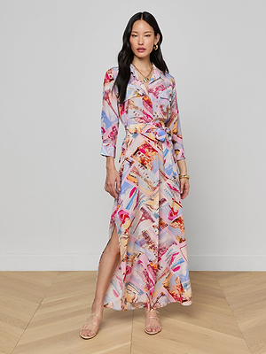 A woman with long dark hair stands against a plain wall wearing the Cameron Silk Shirt Dress, featuring long sleeves and a colorful postcard print in pink, red, blue, and beige. She completes the look with sandals.