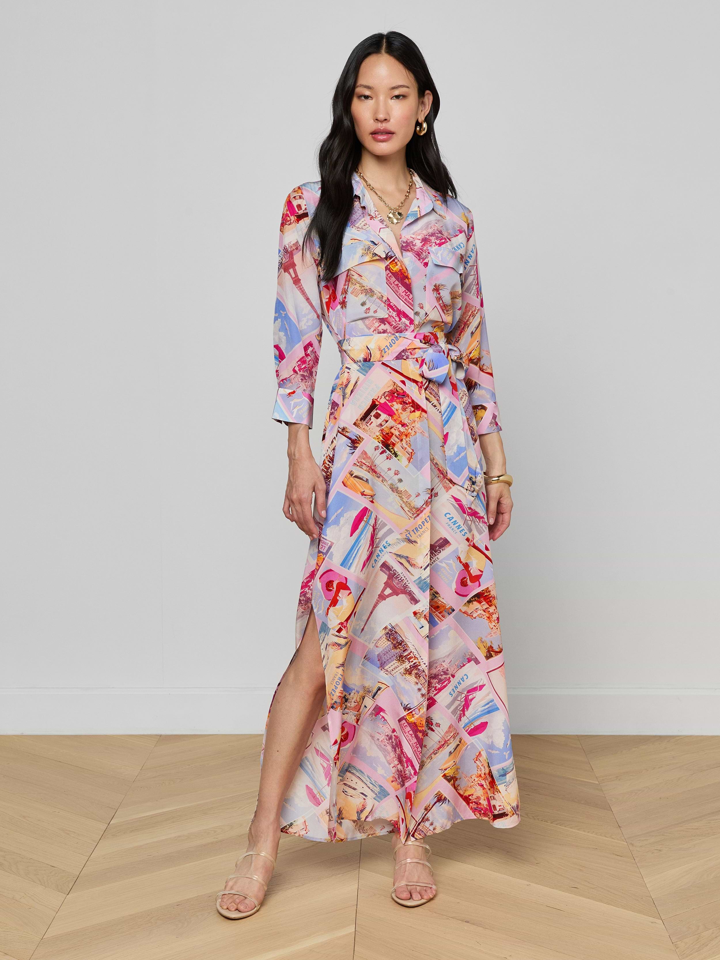 A woman stands wearing the Cameron Silk Shirt Dress, a colorful crepe de chine maxi with long sleeves, a collar, and a side slit. The dress showcases a vibrant abstract pattern in red, pink, blue, and beige tones. She pairs it with sandals, has long black hair, and maintains a neutral expression.