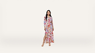 A woman stands wearing a long, colorful, patterned wrap dress with a high slit and long sleeves. She has long hair and wears strappy sandals against a plain white background.