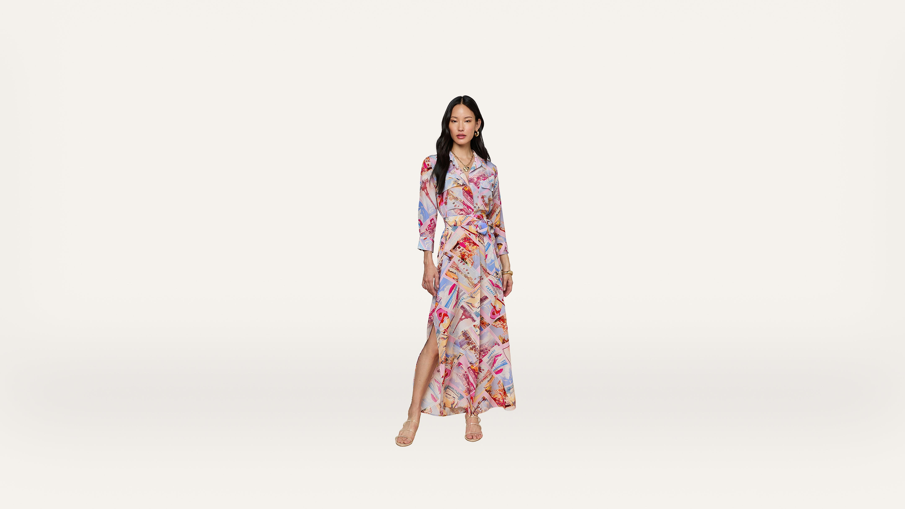 A woman stands wearing a long, colorful, patterned wrap dress with a high slit and long sleeves. She has long hair and wears strappy sandals against a plain white background.