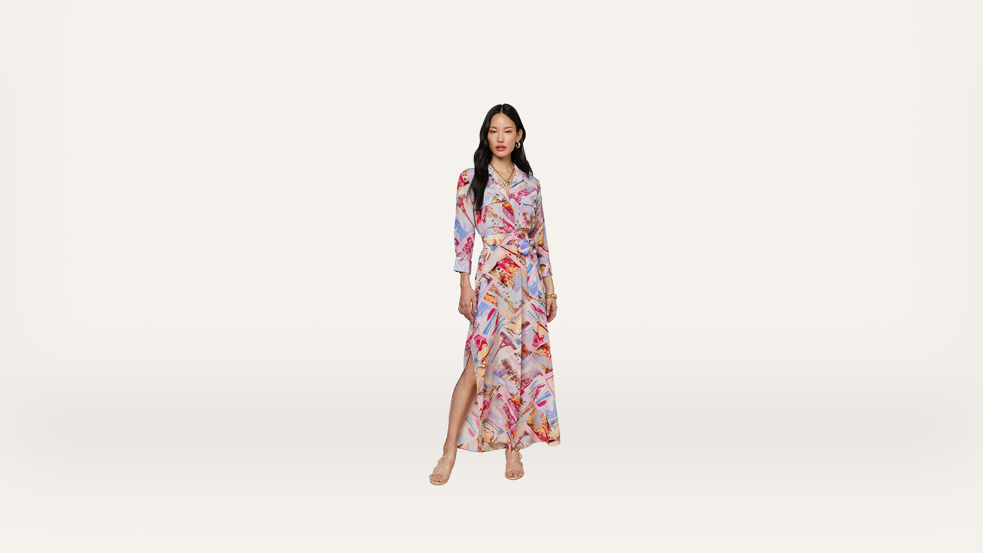 A woman stands wearing a long, colorful, patterned wrap dress with a high slit and long sleeves. She has long hair and wears strappy sandals against a plain white background.