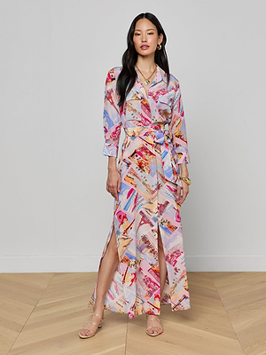 A woman stands indoors wearing the Cameron Silk Shirt Dress, a long silk crepe de chine piece with a colorful postcard print, belted waist, and front slit. She accessorizes with gold jewelry and strappy sandals against a light gray wall and wooden floor.