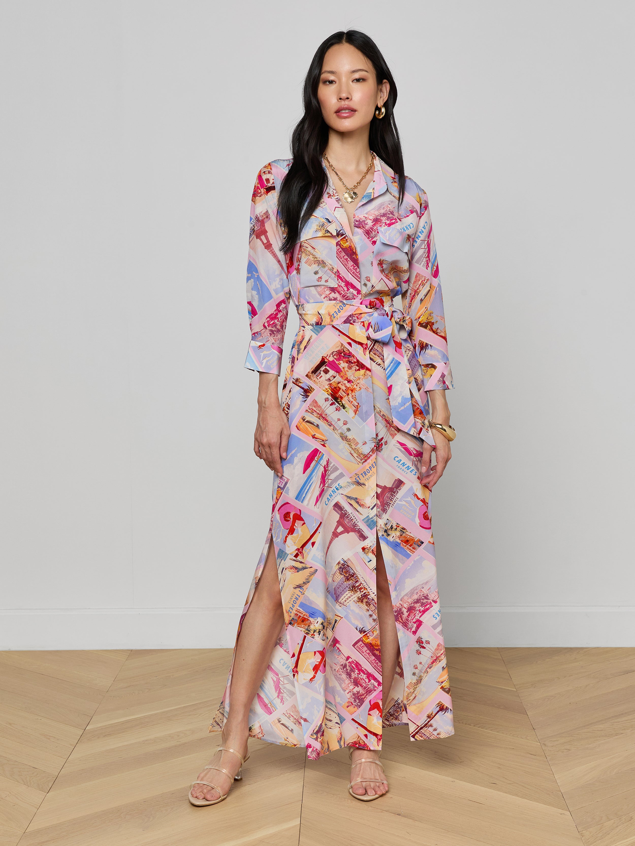 A woman stands against a plain white background wearing the Cameron Silk Shirt Dress, featuring colorful, long sleeves, an ankle-length silhouette, a tied waist, and an abstract postcard print pattern.