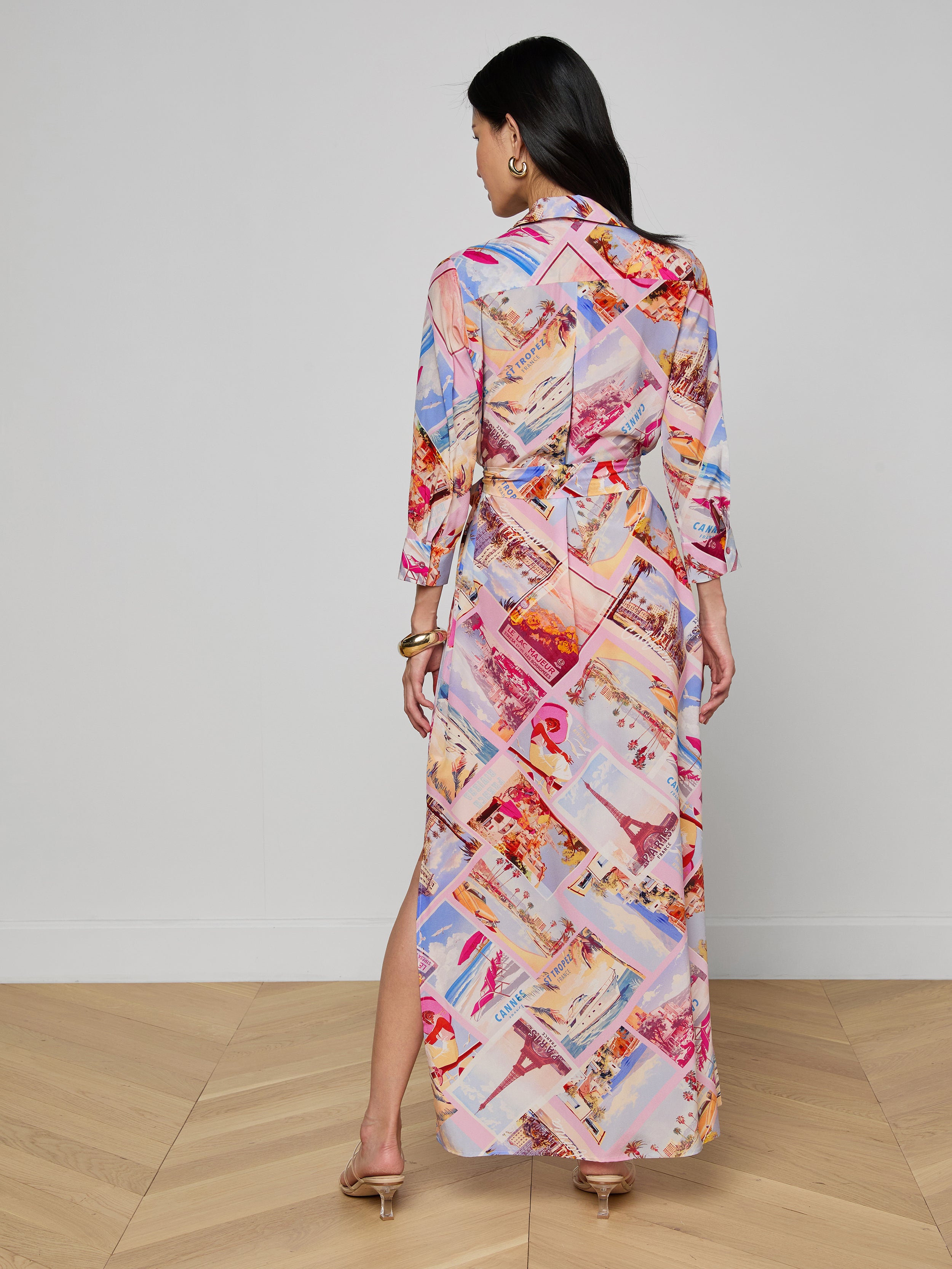 A woman stands in heels, facing away, wearing the Cameron Silk Shirt Dress—a colorful silk crepe de chine maxi dress with a collage-like postcard print and a side slit—against a plain white background.