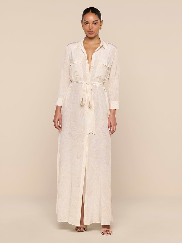 A woman stands against a beige background wearing the Cameron Silk Shirt Dress—a long, light beige piece with buttoned cuffs, chest pockets, a front slit, and a subtle paisley pattern. Her hair is pulled back and she pairs it with sandals.