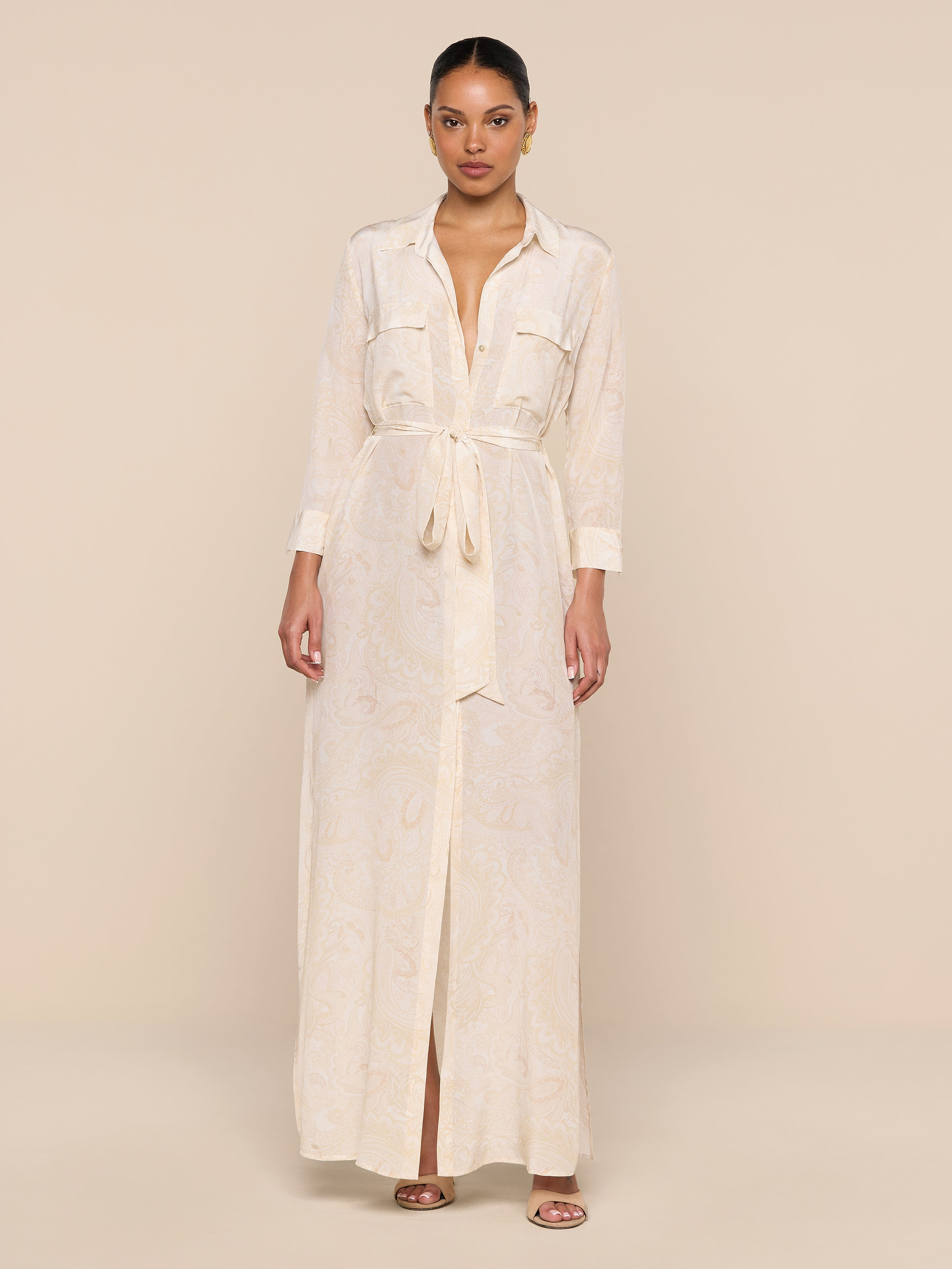 A woman stands against a beige background wearing the Cameron Silk Shirt Dress—a long, light beige piece with buttoned cuffs, chest pockets, a front slit, and a subtle paisley pattern. Her hair is pulled back and she pairs it with sandals.
