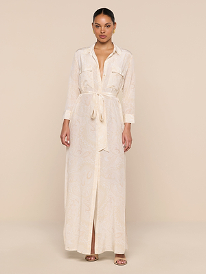 A woman stands against a beige background wearing the Cameron Silk Shirt Dress—a long, light beige piece with buttoned cuffs, chest pockets, a front slit, and a subtle paisley pattern. Her hair is pulled back and she pairs it with sandals.