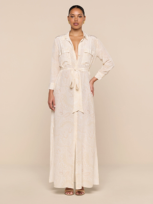 A woman stands against a beige background wearing the Cameron Silk Shirt Dress—a long paisley print dress with long sleeves, front pockets, and a tie belt at the waist—styled with open-toe sandals.
