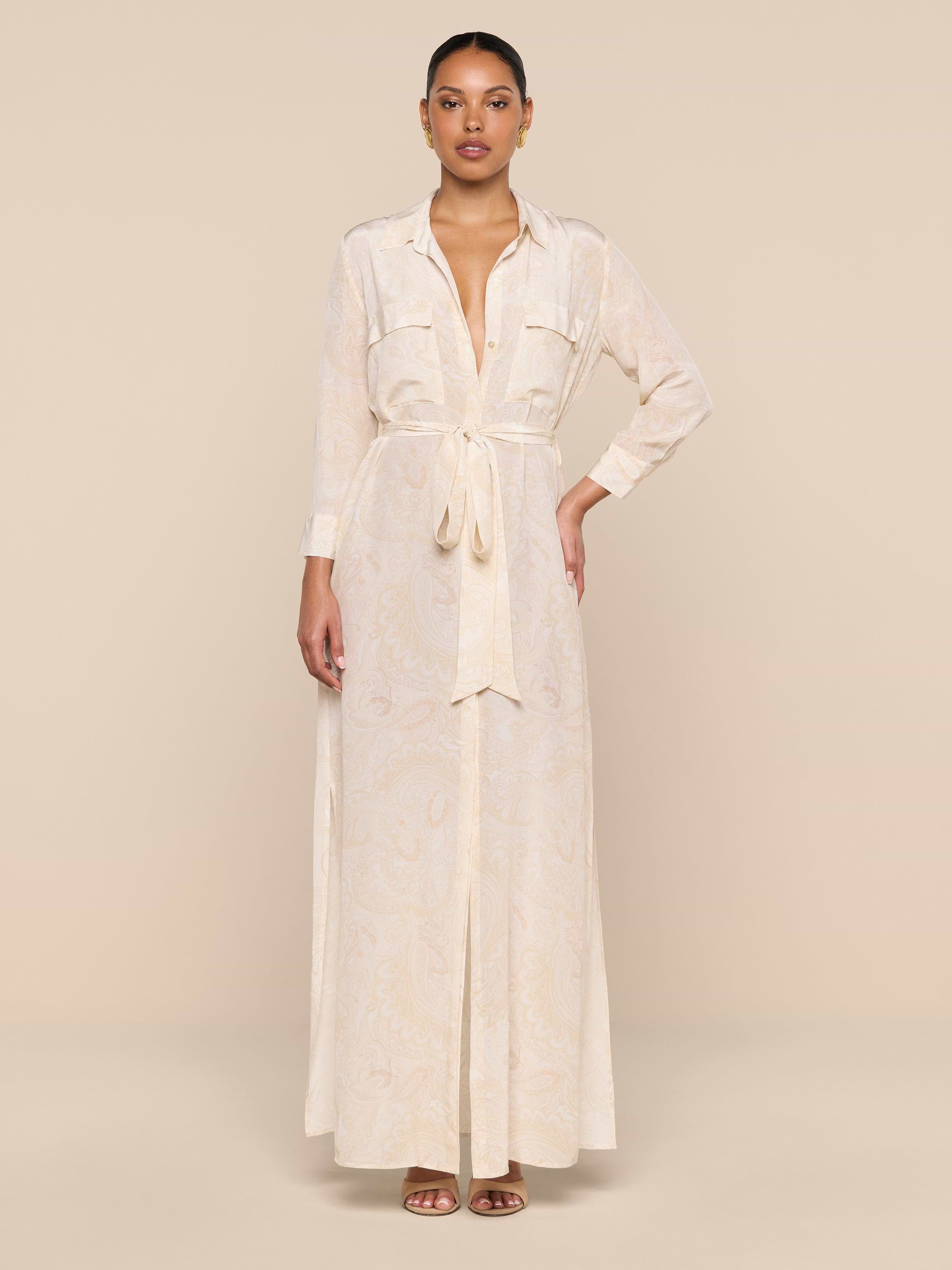 A woman stands against a beige background wearing the Cameron Silk Shirt Dress—a long paisley print dress with long sleeves, front pockets, and a tie belt at the waist—styled with open-toe sandals.