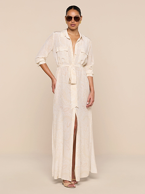 A woman poses against a beige background in the Cameron Silk Shirt Dress—a long, light silk piece with a subtle paisley print, belted waist, rolled sleeves—paired with sunglasses and heeled sandals. One hand rests on her hip.