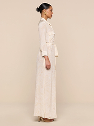 A woman in profile stands against a beige background, wearing the Cameron Silk Shirt Dress with long sleeves and pockets, gold hoop earrings, brown heeled sandals, and her hair styled in a neat bun.