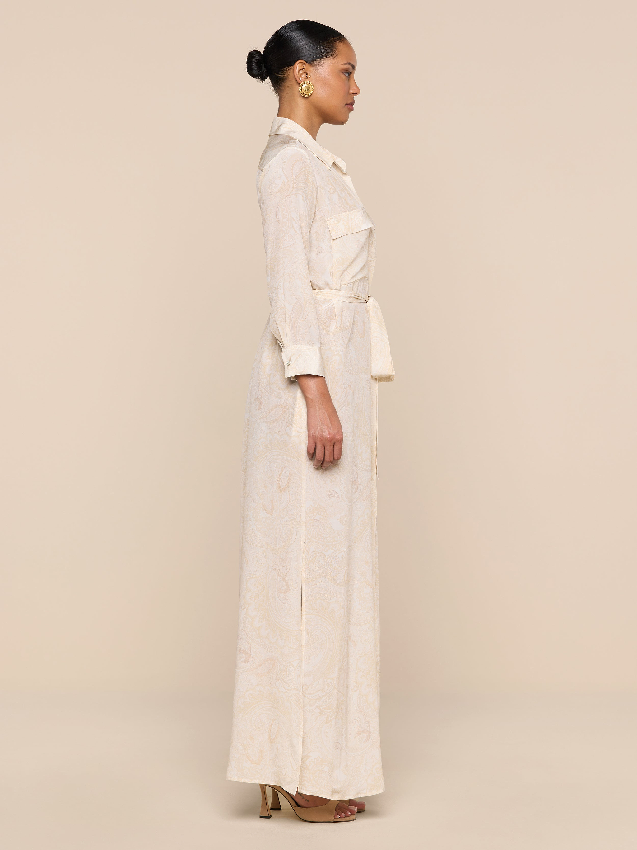 A woman in profile stands against a beige background, wearing the Cameron Silk Shirt Dress with long sleeves and pockets, gold hoop earrings, brown heeled sandals, and her hair styled in a neat bun.