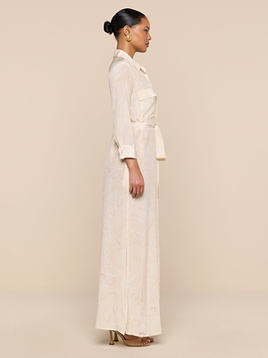 A woman in profile stands against a beige background, wearing the Cameron Silk Shirt Dress with long sleeves and pockets, gold hoop earrings, brown heeled sandals, and her hair styled in a neat bun.