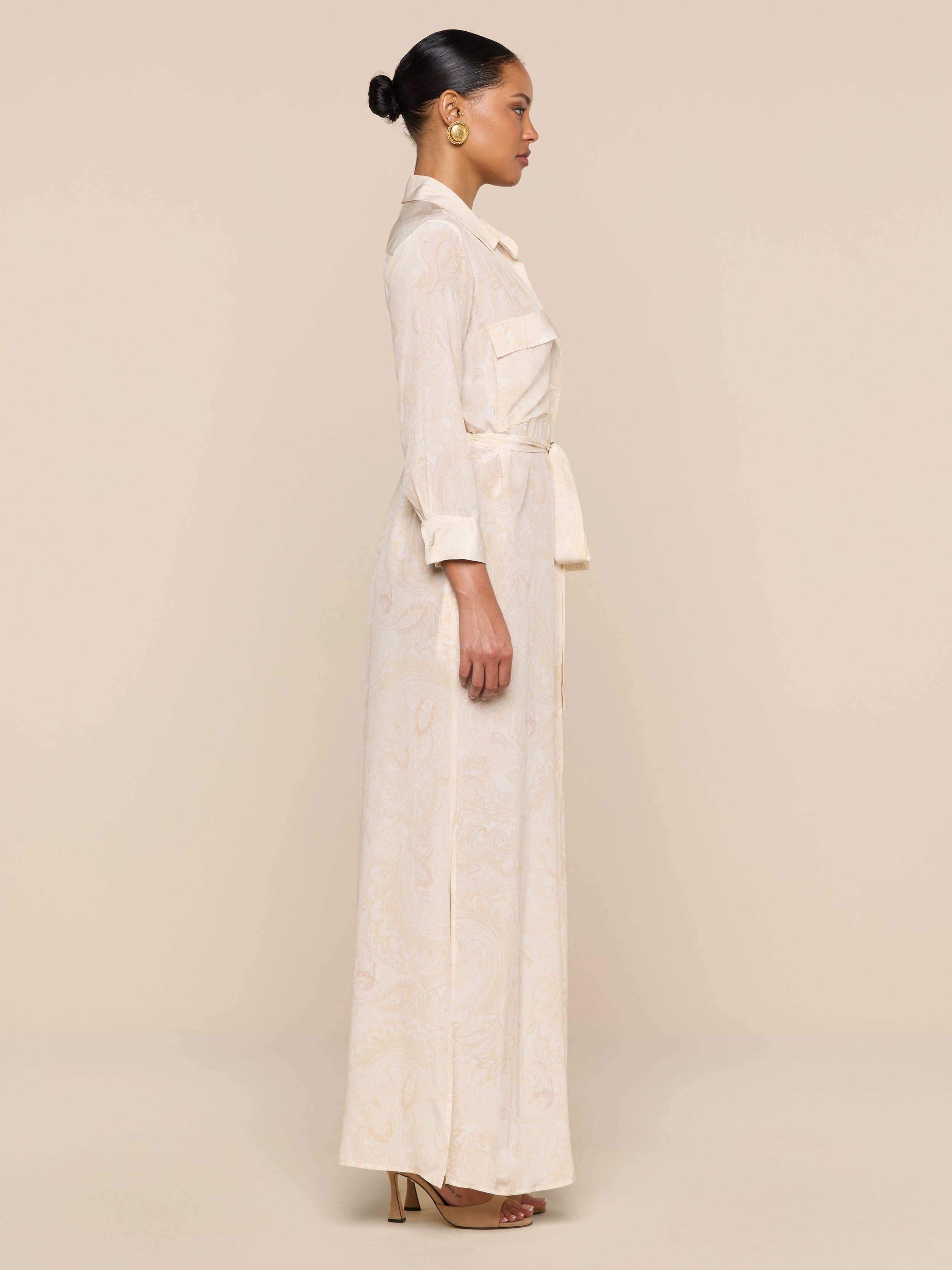 A woman in profile stands against a beige background, wearing the Cameron Silk Shirt Dress with long sleeves and pockets, gold hoop earrings, brown heeled sandals, and her hair styled in a neat bun.