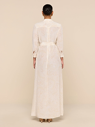 A woman with her hair in a bun models the Cameron Silk Shirt Dress—long, cream-colored, belted, with long sleeves—while standing with her back to the camera against a beige background.