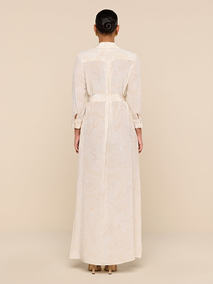A woman with her hair in a bun models the Cameron Silk Shirt Dress—long, cream-colored, belted, with long sleeves—while standing with her back to the camera against a beige background.