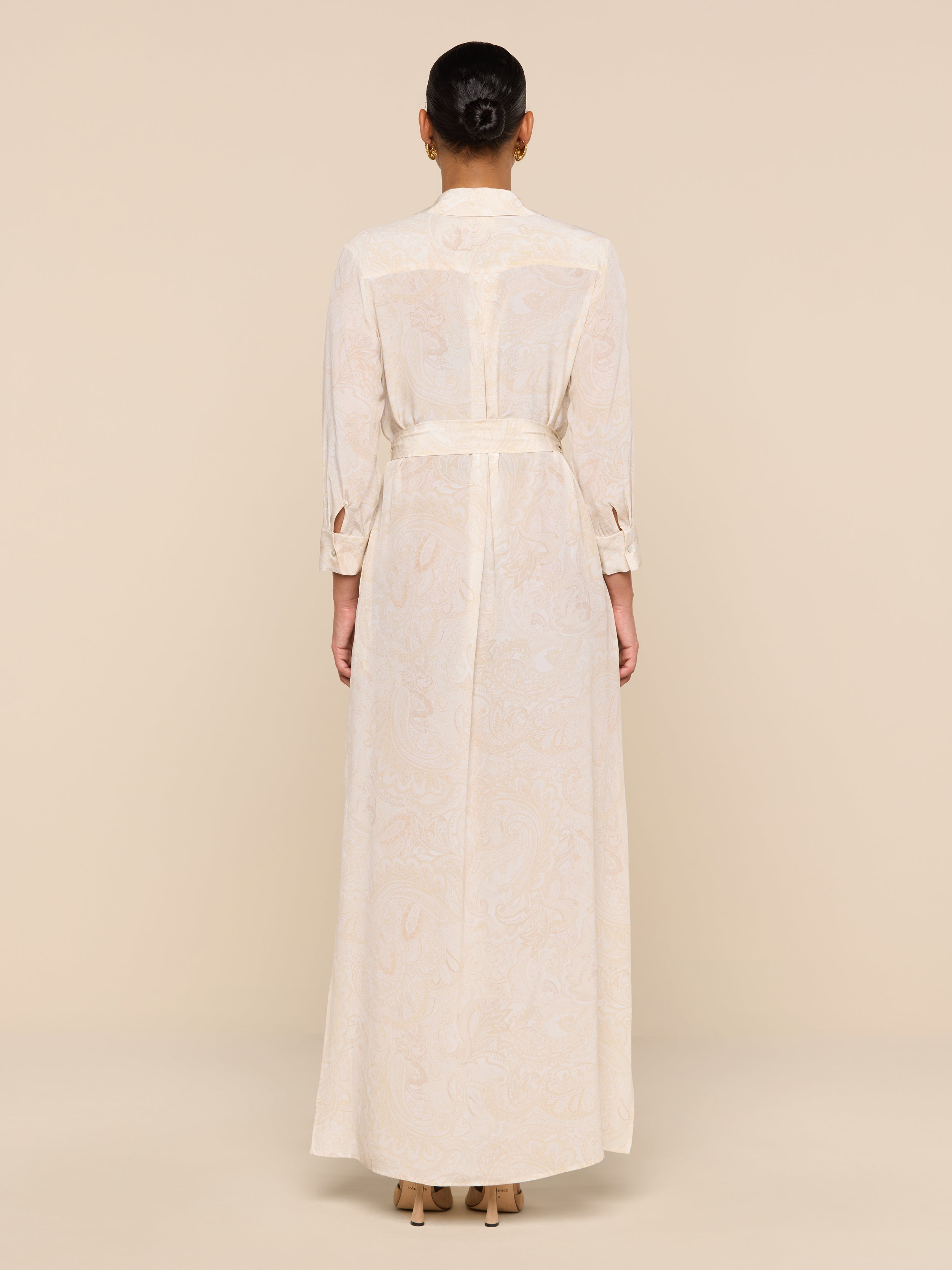 A woman with her hair in a bun models the Cameron Silk Shirt Dress—long, cream-colored, belted, with long sleeves—while standing with her back to the camera against a beige background.