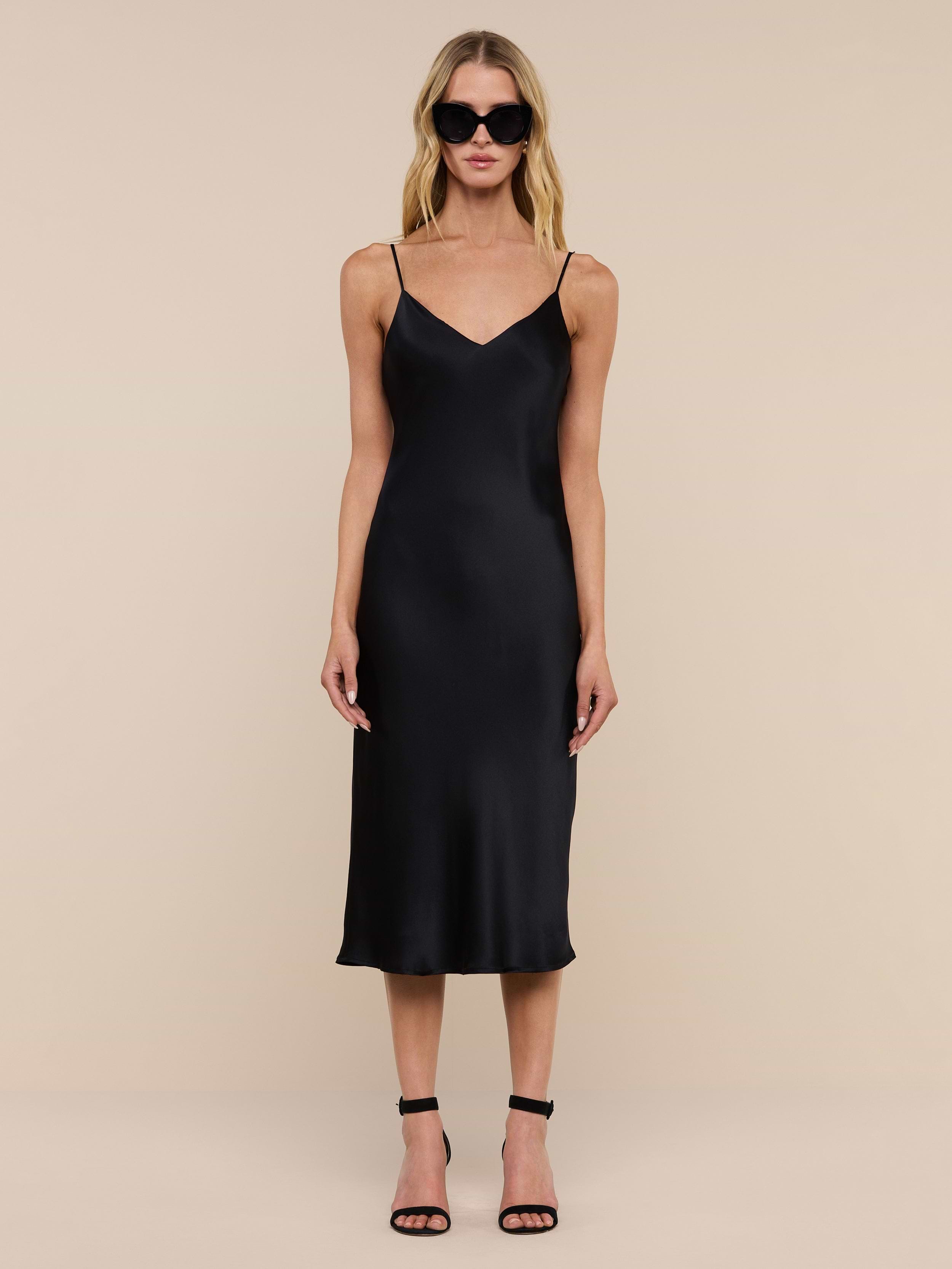 A woman wearing the Jodie Silk Slip Dress in black, paired with black high-heeled sandals and large dark sunglasses, stands against a plain white background, facing forward with her arms relaxed at her sides.