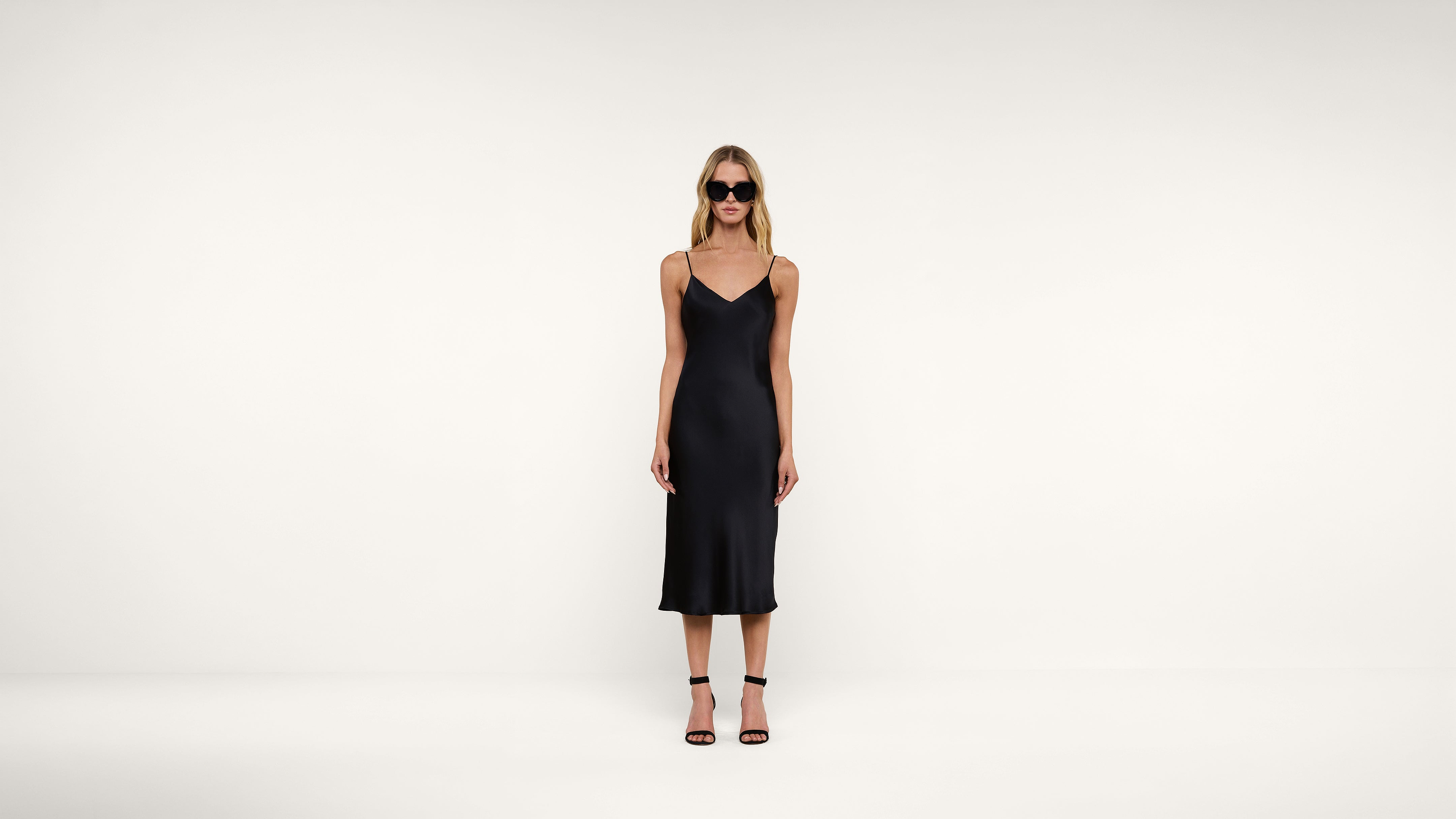 Woman wearing a black, sleeveless, midi slip dress with thin straps, black heels, and sunglasses, standing against a plain white background.