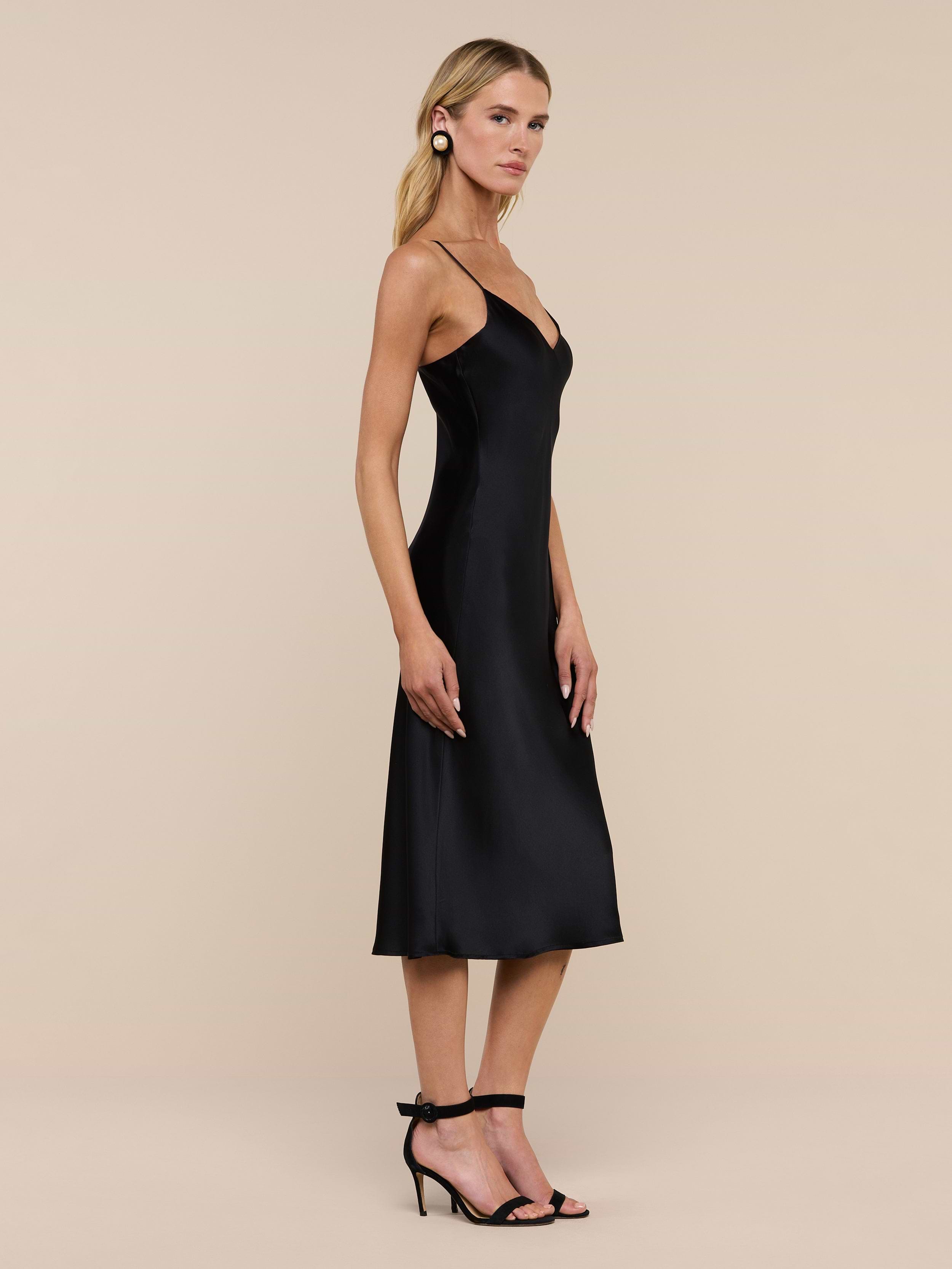 A woman with long blonde hair and hoop earrings stands sideways against a plain white background, wearing the Jodie Silk Slip Dress in black paired with black heels.