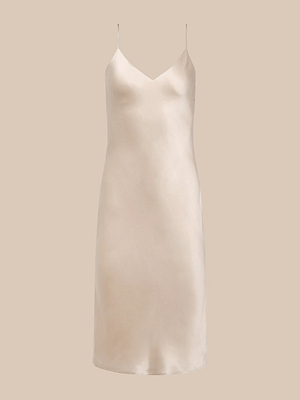 The Jodie Silk Slip Dress is a knee-length, sleeveless champagne slip dress in light beige satin, featuring thin spaghetti straps and a V-neckline, shown on a plain beige background.