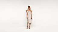 A woman stands against a plain white background, wearing a light-colored, sleeveless midi dress and strappy heels. She faces forward with a neutral expression and her hair styled in loose waves.