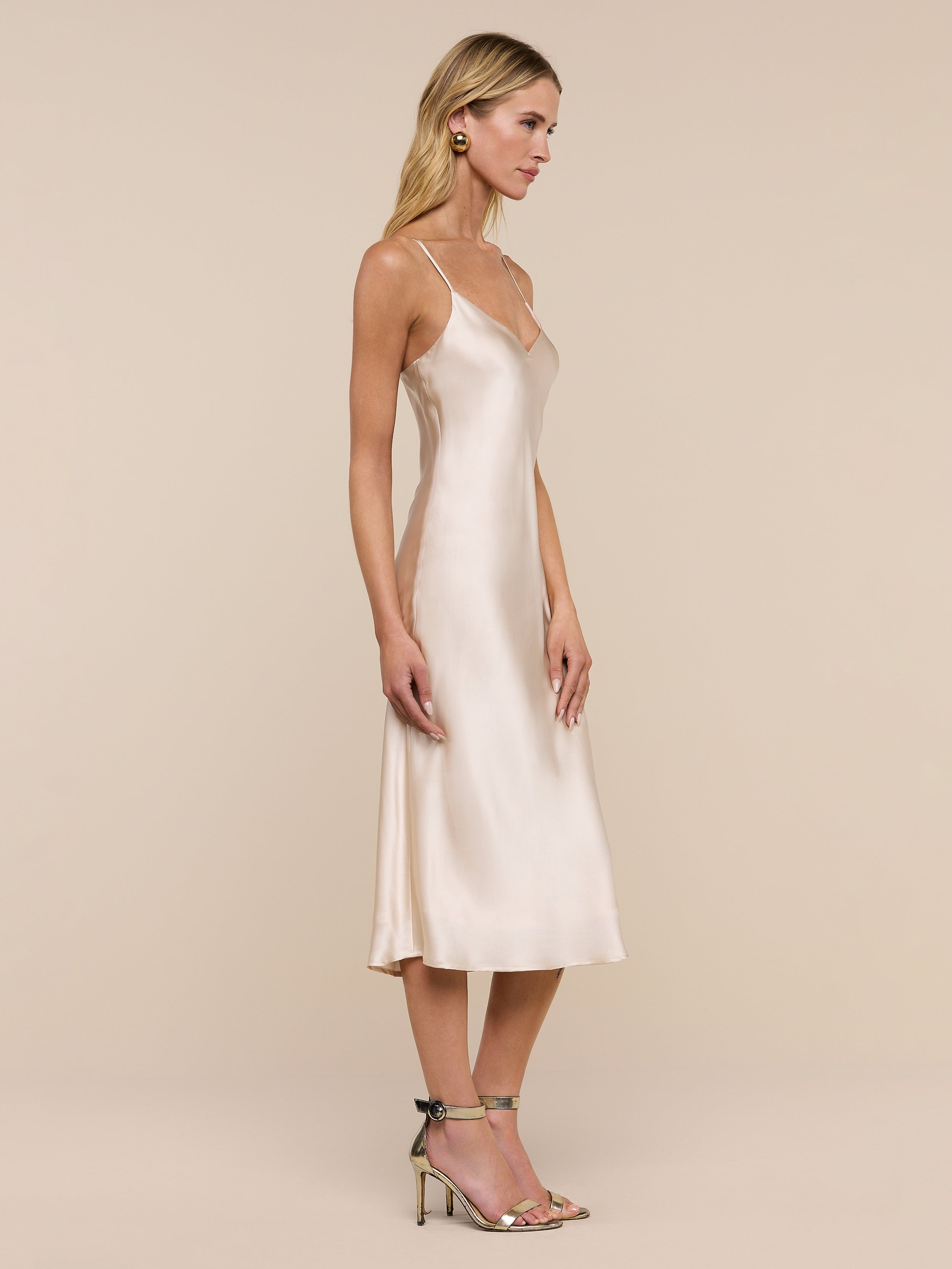 A woman wearing the Jodie Silk Slip Dress, a knee-length silk charmeuse design, and heeled sandals stands in profile against a plain white background.