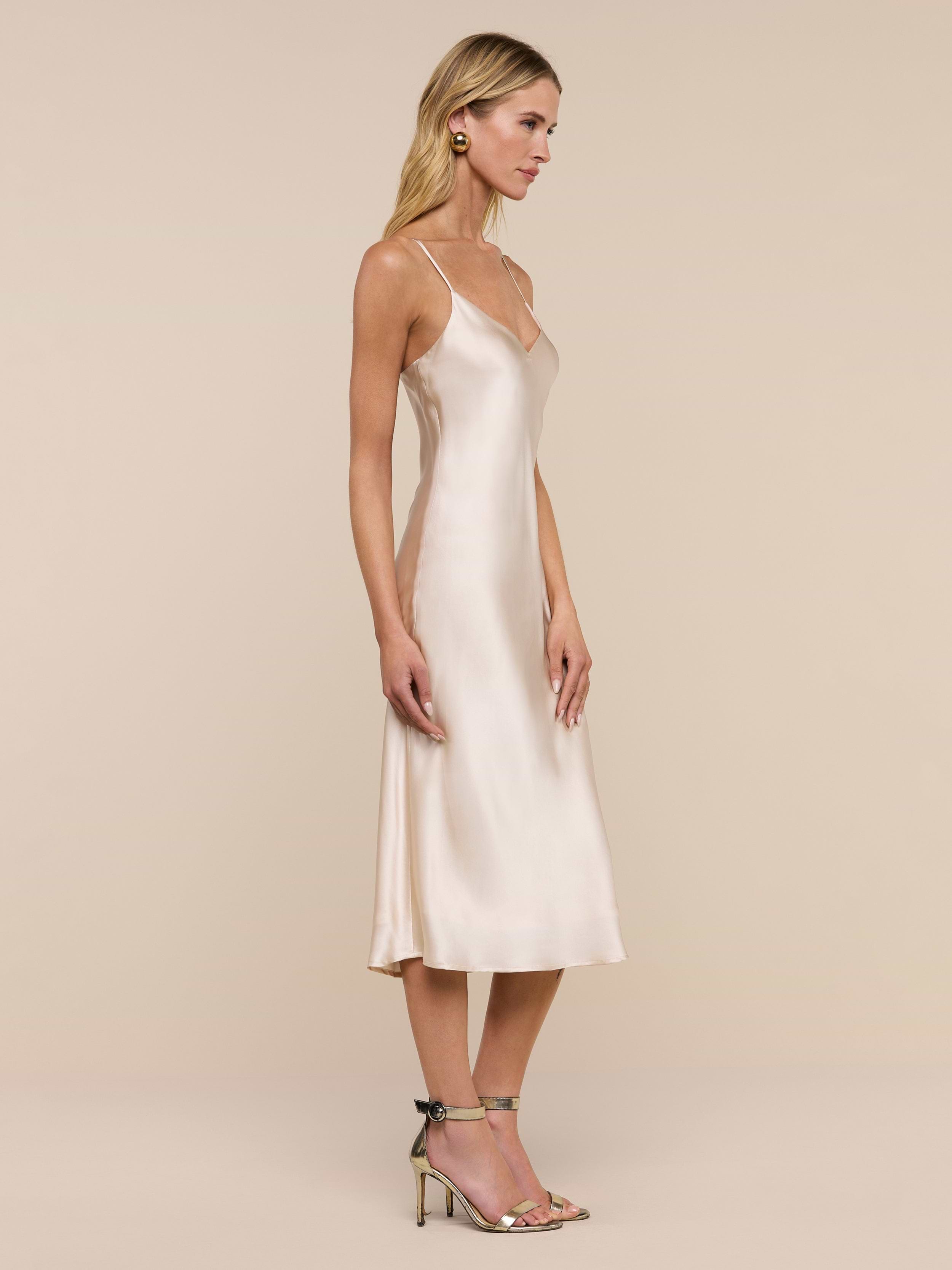 A woman wearing the Jodie Silk Slip Dress, a knee-length silk charmeuse design, and heeled sandals stands in profile against a plain white background.