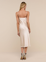 A person with long blond hair, standing and facing away from the camera, is wearing the Jodie Silk Slip Dress in champagne silk charmeuse with high-heeled shoes against a plain white background.