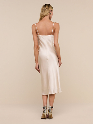 A woman faces away, wearing the Jodie Silk Slip Dress in light champagne with thin straps and midi length. Her long, wavy blonde hair and silver ankle-strap heels enhance the elegant satin look against a plain beige background.
