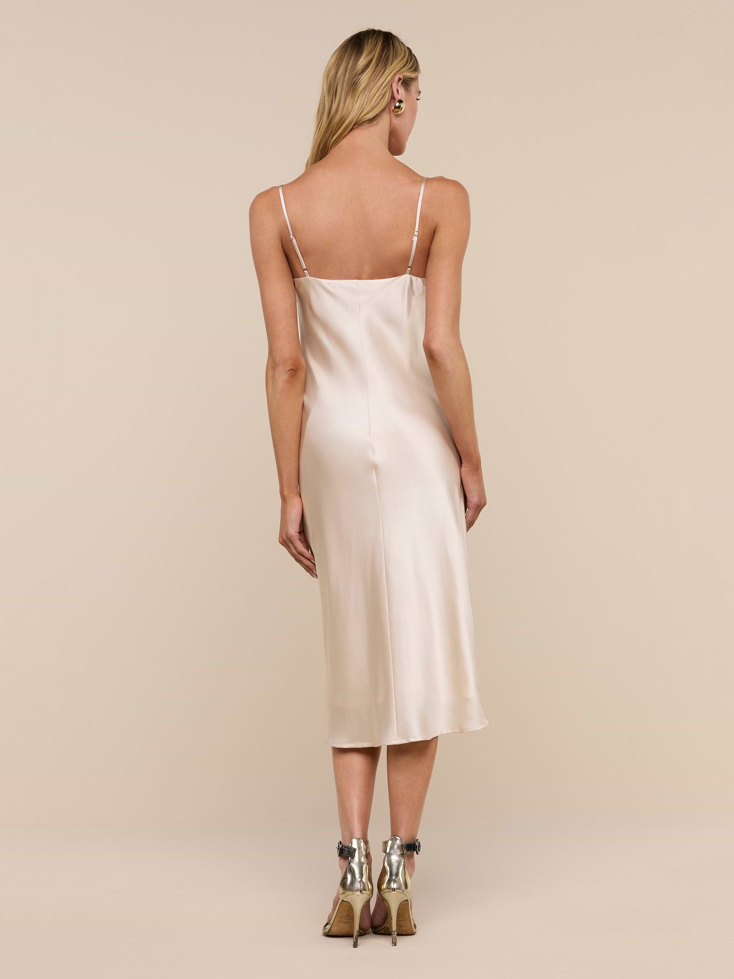 A person with long blond hair, standing and facing away from the camera, is wearing the Jodie Silk Slip Dress in champagne silk charmeuse with high-heeled shoes against a plain white background.