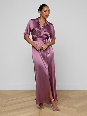 A woman wears the Klement Silk Shirt Dress, a mauve short-sleeve style with a tie waist, cargo pockets, and front slit. She pairs it with silver heels; her hair is pulled back. The plain background features light wood flooring.
