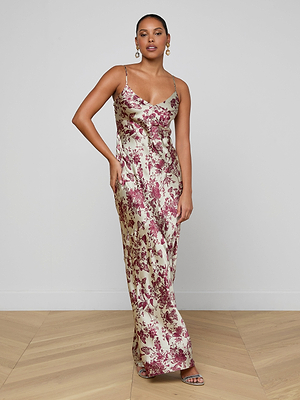 A woman stands on a wooden floor wearing the Serita Silk Gown, a long, sleeveless bias-cut dress with thin straps and a pink floral butterfly print on cream. She finishes the look with hoop earrings and open-toe heels.