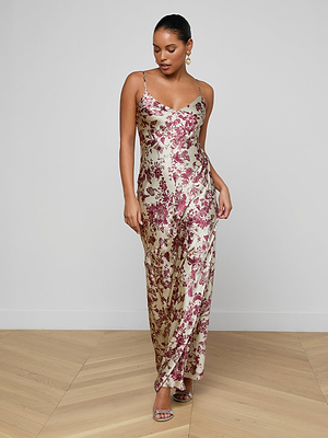 A woman stands on a wooden floor against a white wall, wearing the Serita Silk Gown—a long gold slip dress with a pink floral butterfly print. She pairs it with hoop earrings, strappy heels, and has her hair pulled back.