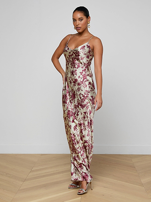 A woman poses elegantly in a minimalist room wearing the Serita Silk Gown, a long cream-colored slip dress with a red floral butterfly print. Her hair is pulled back, and she accessorizes with earrings and strappy heels.
