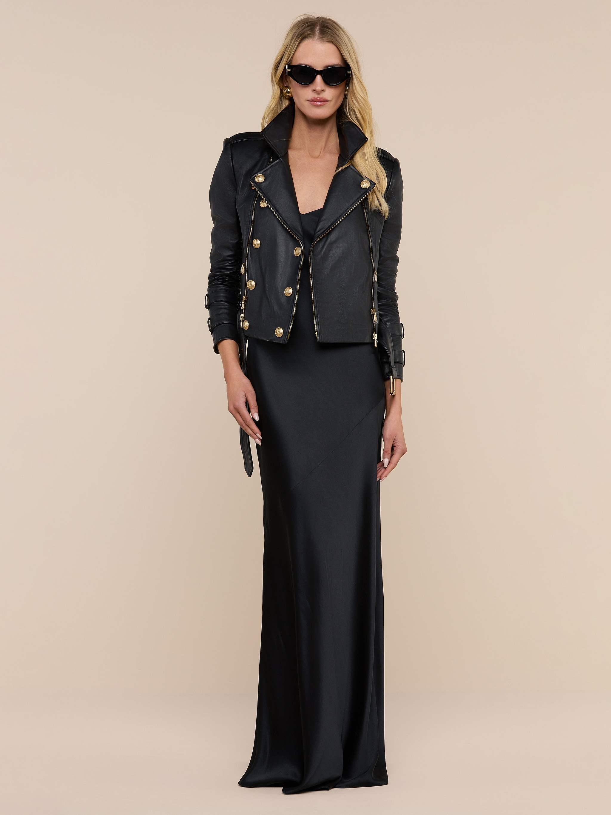A woman wearing the Serita Silk Gown—a long black silk dress—paired with a black leather jacket featuring gold buttons and black sunglasses stands against a plain white background.