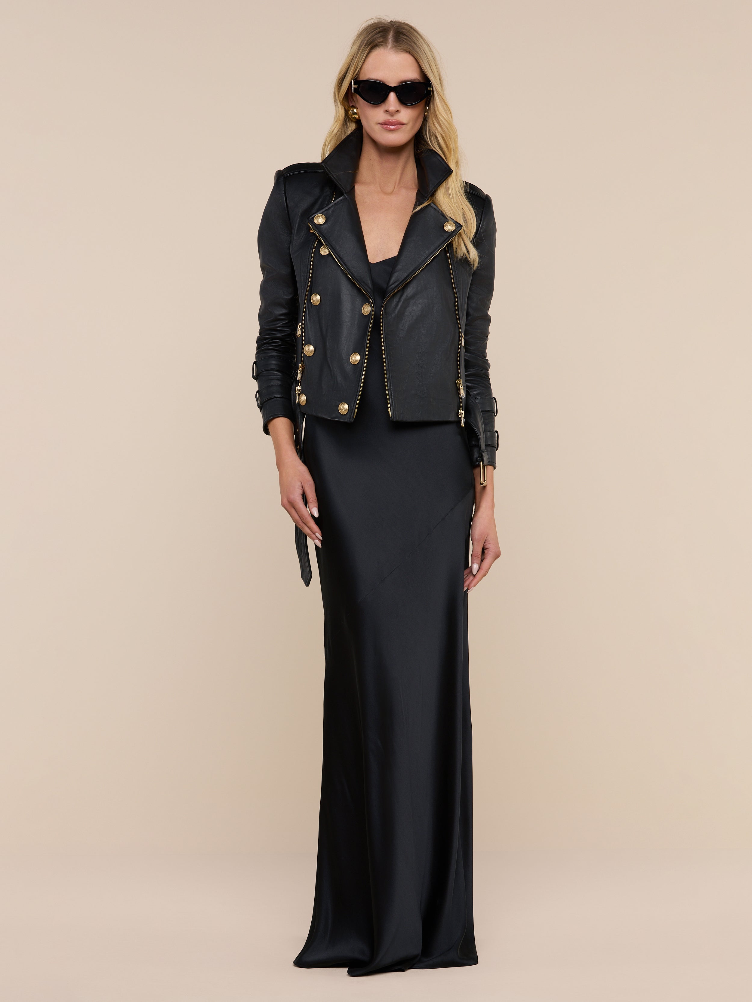 A woman wearing the Serita Silk Gown—a long black silk dress—paired with a black leather jacket featuring gold buttons and black sunglasses stands against a plain white background.