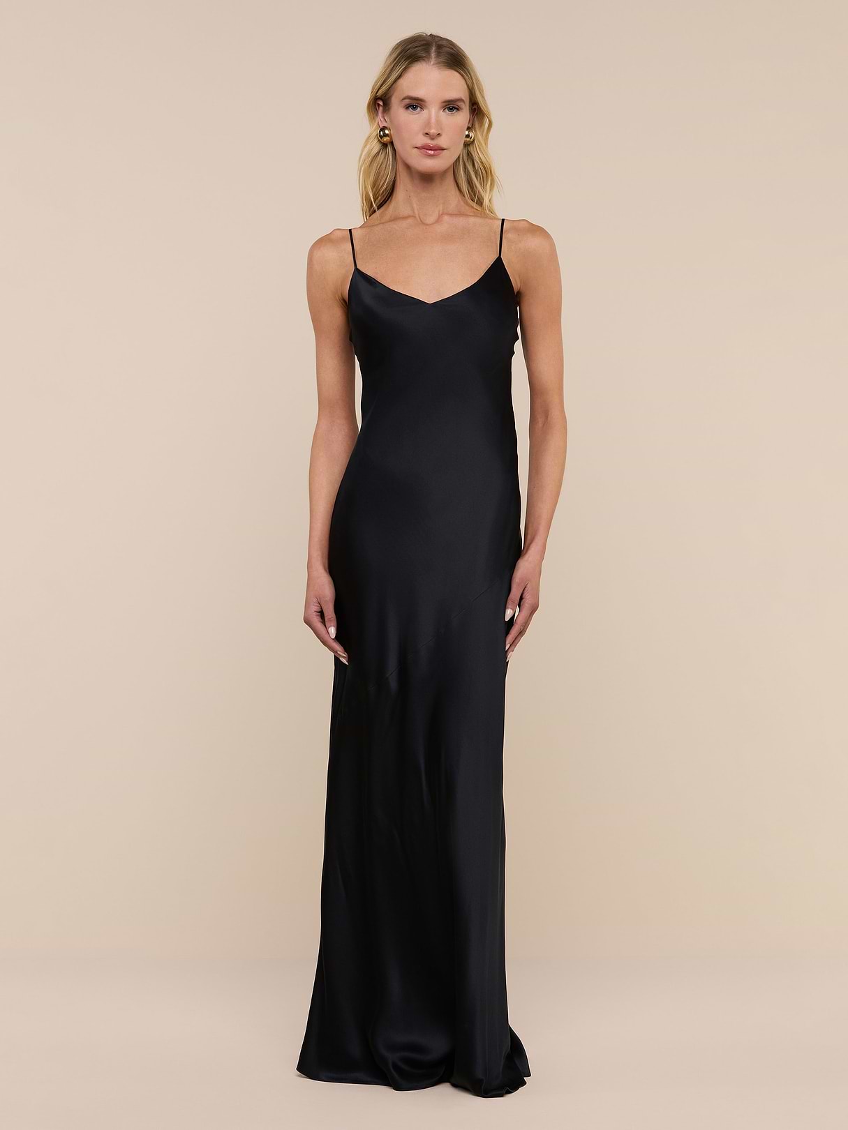 A woman stands against a plain white background wearing the Serita Silk Gown—a long, sleeveless black dress with thin straps, crafted from luxurious silk charmeuse.