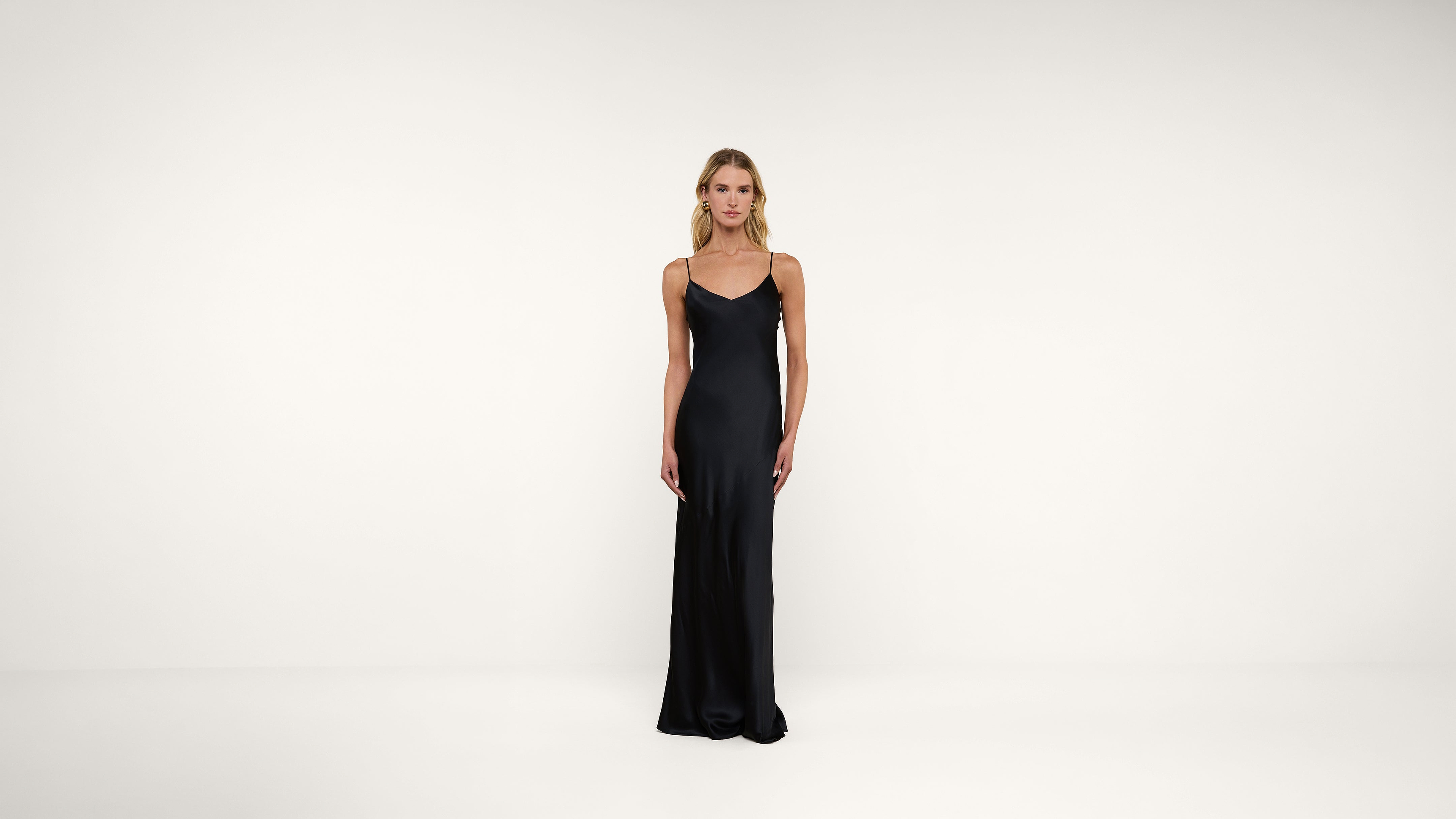 A woman stands against a plain white background, wearing a long, black, sleeveless slip dress with thin straps.