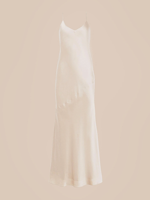 The Serita Silk Gown is a long, sleeveless champagne slip dress with thin spaghetti straps, a gentle V-neckline, and a subtle sheen, shown against a plain beige background.