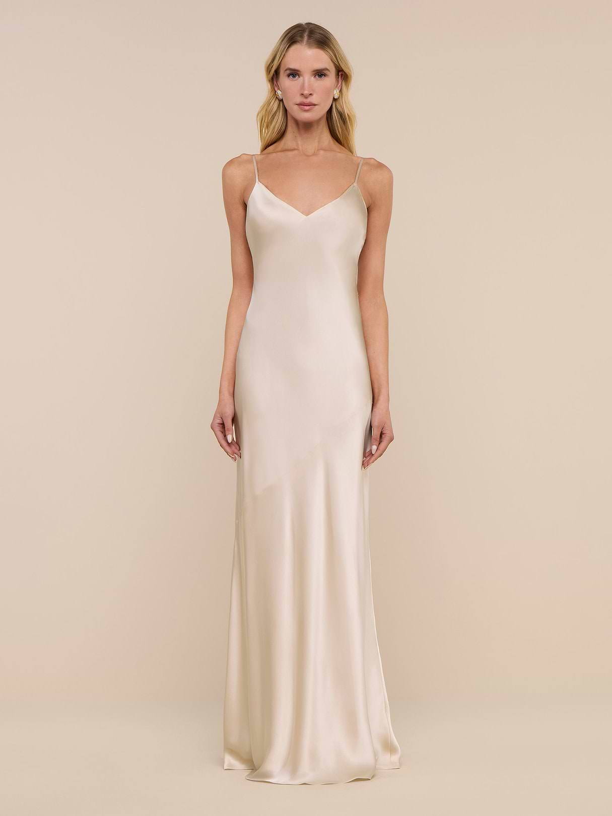 A woman stands against a plain white background, wearing the Serita Silk Gown—a long, sleeveless charmeuse slip dress in a champagne hue with thin straps.