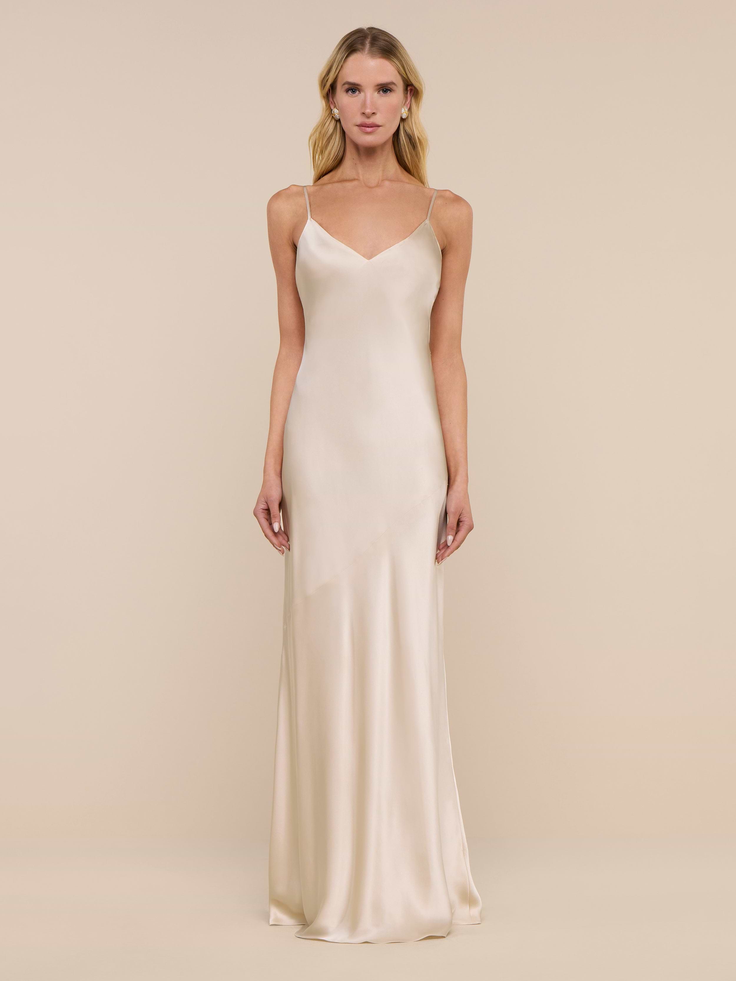 A woman stands against a plain white background, wearing the Serita Silk Gown—a long, sleeveless charmeuse slip dress in a champagne hue with thin straps.