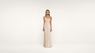 A woman stands against a plain white background wearing a long, sleeveless, light beige satin dress with thin straps and a simple, elegant design.