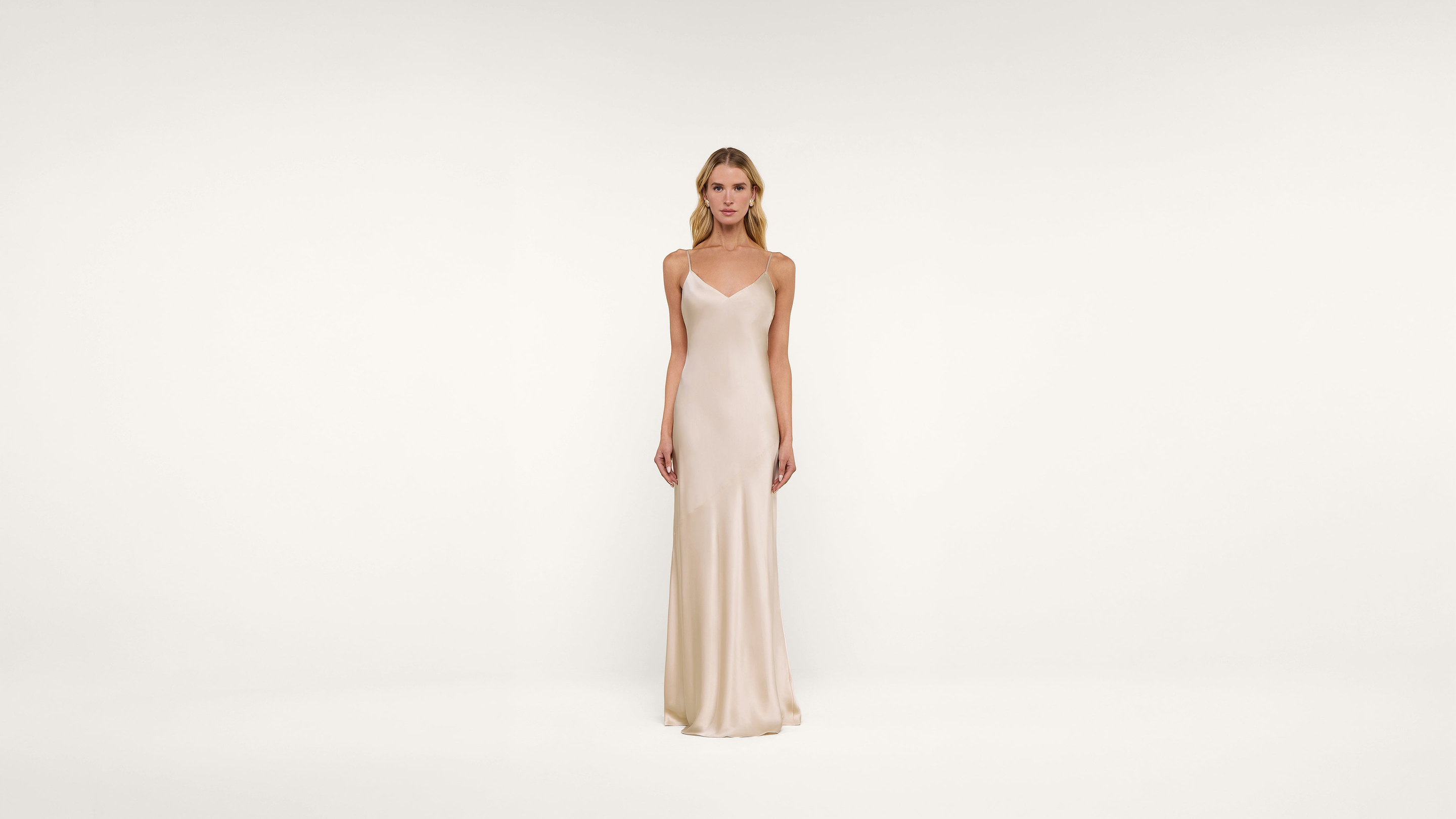 A woman stands against a plain white background wearing a long, sleeveless, light beige satin dress with thin straps and a simple, elegant design.