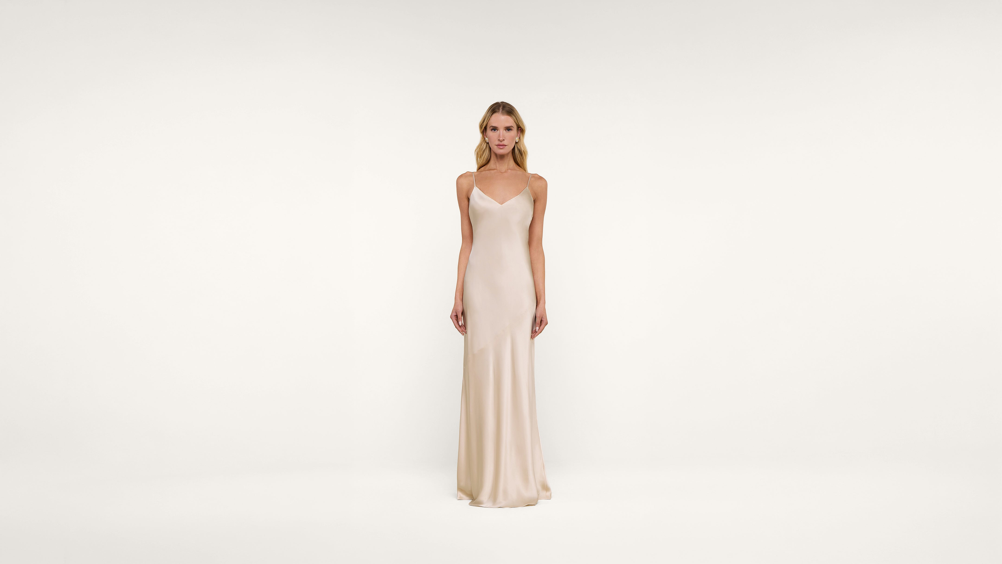 A woman stands against a plain white background wearing a long, sleeveless, light beige satin dress with thin straps and a simple, elegant design.