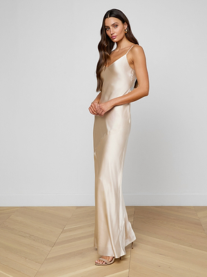 A woman with long brown hair wears the Serita Silk Gown in shiny champagne silk charmeuse and nude sandals, standing on a light wooden floor against a plain white wall.